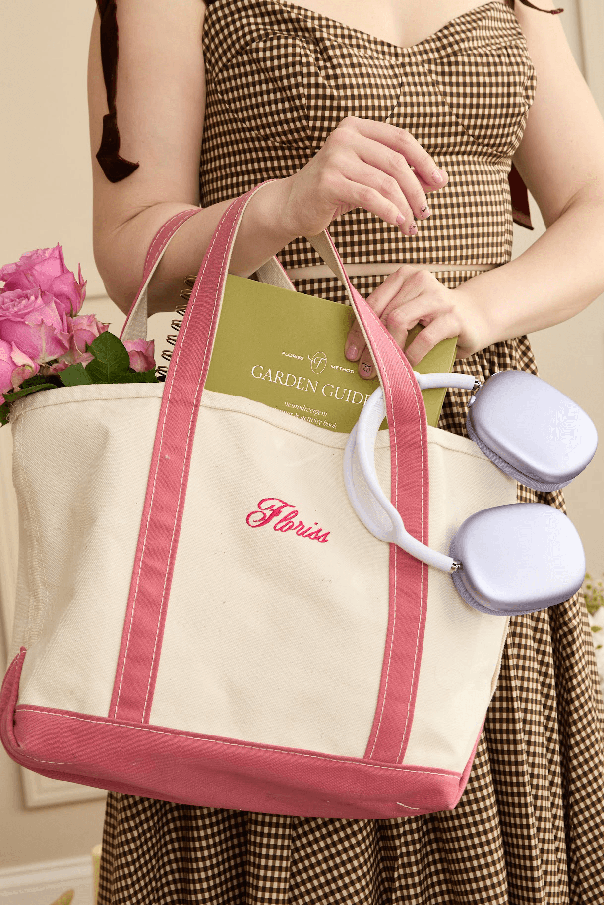 A woman in a checkered brown gingham dress holds a cream and pink tote bag embroidered with "Floriss." The bag contains pink roses, a Floriss Garden Guide book, and purple headphones.
