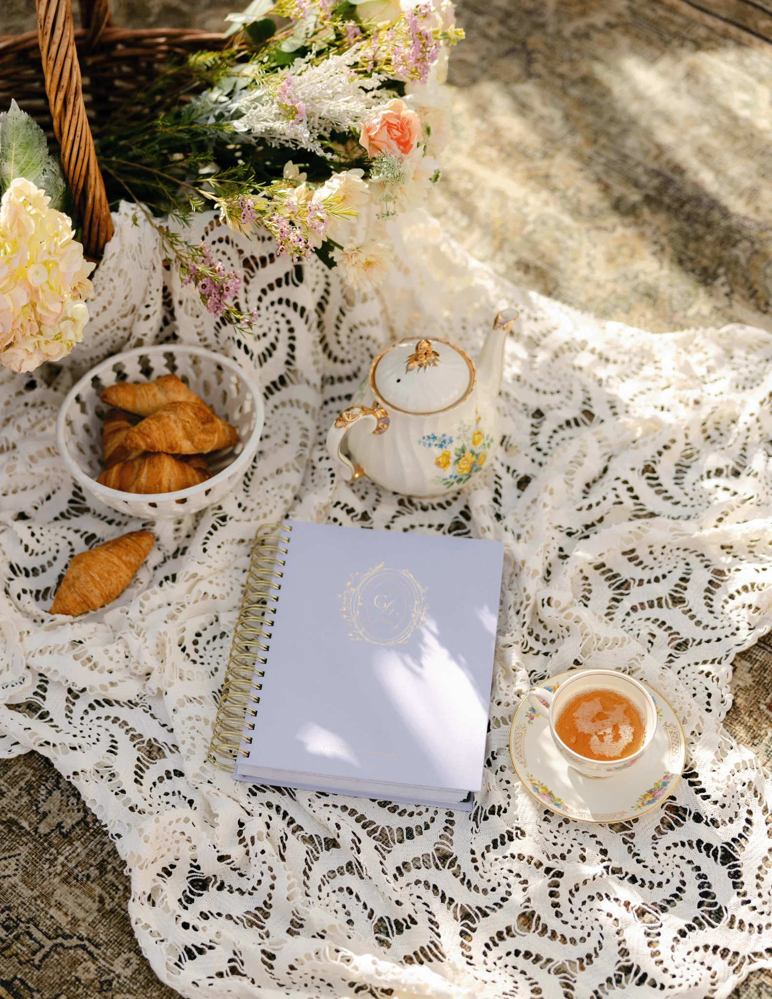 Cozy picnic scene on lace fabric with a teapot, cappuccino in a floral cup and saucer, croissants, a fresh bouquet, and a lavender Floriss Garden Guide planner, representing The Grove Garden Club